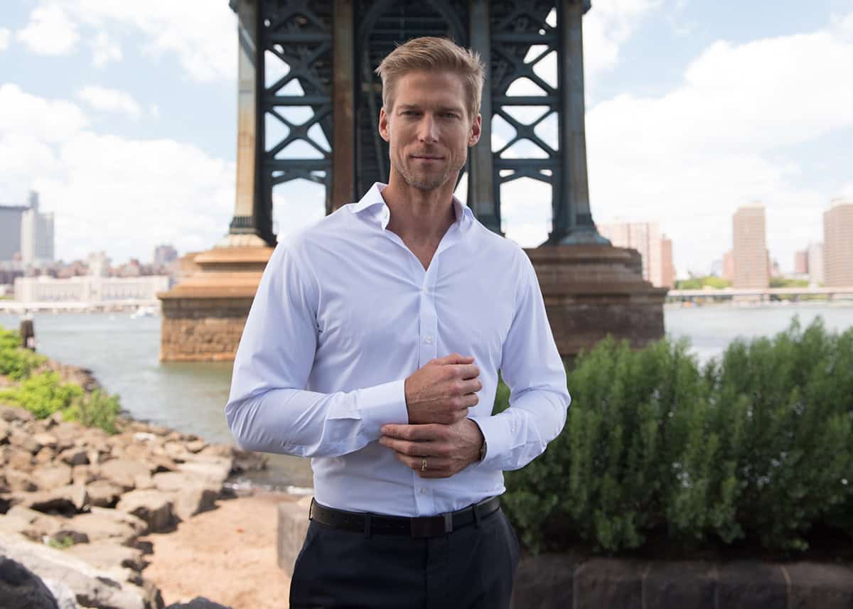 Elegant man in white dress shirt under Brooklyn Bridge, NYC, showcasing confidence and style for DeoVeritas luxury fashion brand.