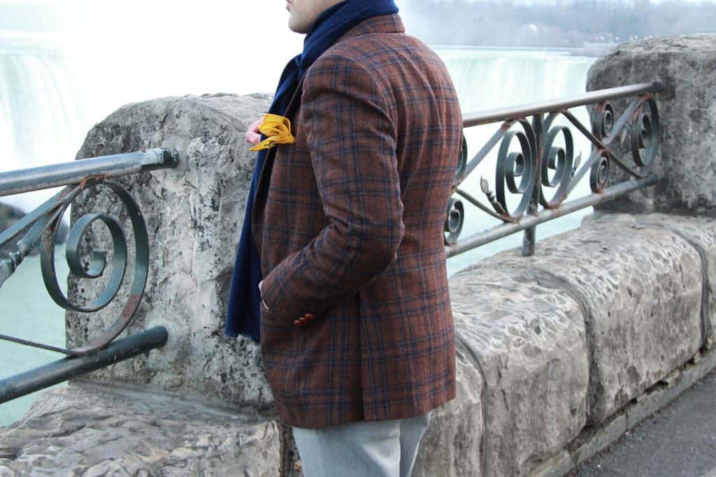 Vivid image of a well-dressed man standing by the railing near Niagara Falls, showcasing stylish winter fashion including a plaid blazer, scarf, and yellow accessory, with iconic cascading waterfalls in the background.