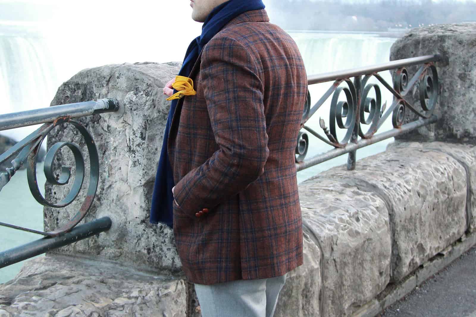 Vivid image of a well-dressed man standing by the railing near Niagara Falls, showcasing stylish winter fashion including a plaid blazer, scarf, and yellow accessory, with iconic cascading waterfalls in the background.