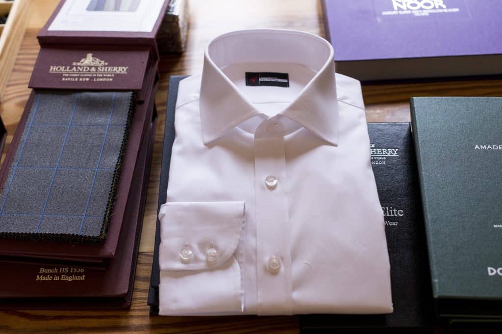 Elegant white dress shirt with classic collar and button details displayed on a wooden surface, surrounded by fabric swatches and brand packaging, emphasizing quality men's formalwear for stylish occasions.