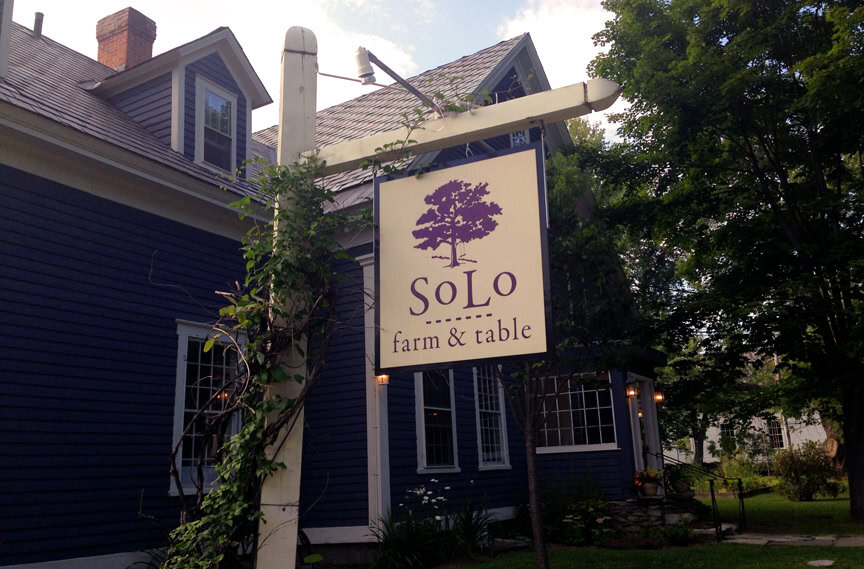 Solo farm and table restaurant signage in an outdoor setting with a blue house and lush green trees, emphasizing farm-to-table dining, local food, fresh ingredients, and rustic farm atmosphere.