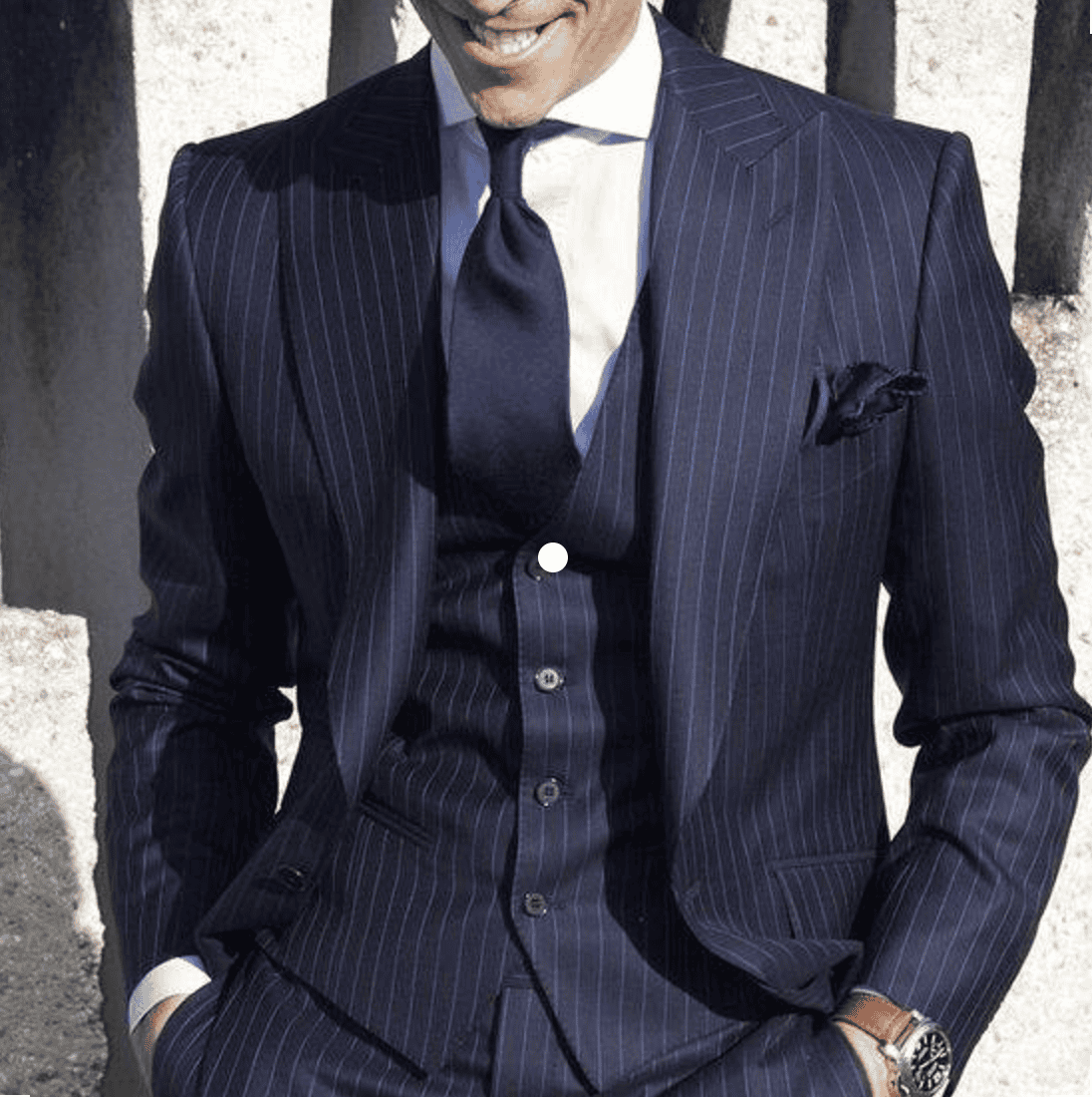 Business man wearing navy blue pinstripe suit with matching tie and pocket square, standing outdoors in front of stone wall, confident professional appearance, luxury fashion, men's formal wear, sophisticated style, DeoVeritas branding.