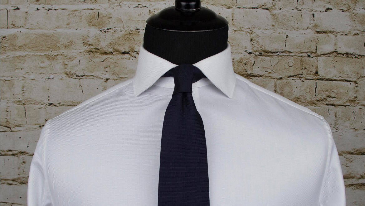 High-quality white dress shirt with a navy tie displayed on a mannequin against a textured brick wall background. Suitable for professional or formal occasions.