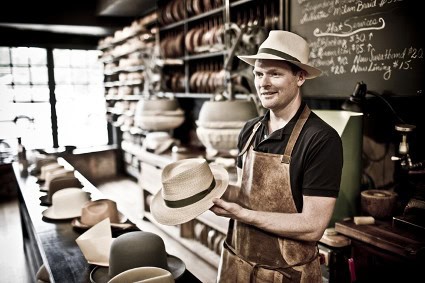 Handsome man in a hat shop holding a stylish Panama hat, surrounded by various hat styles in a cozy, well-lit store setting.