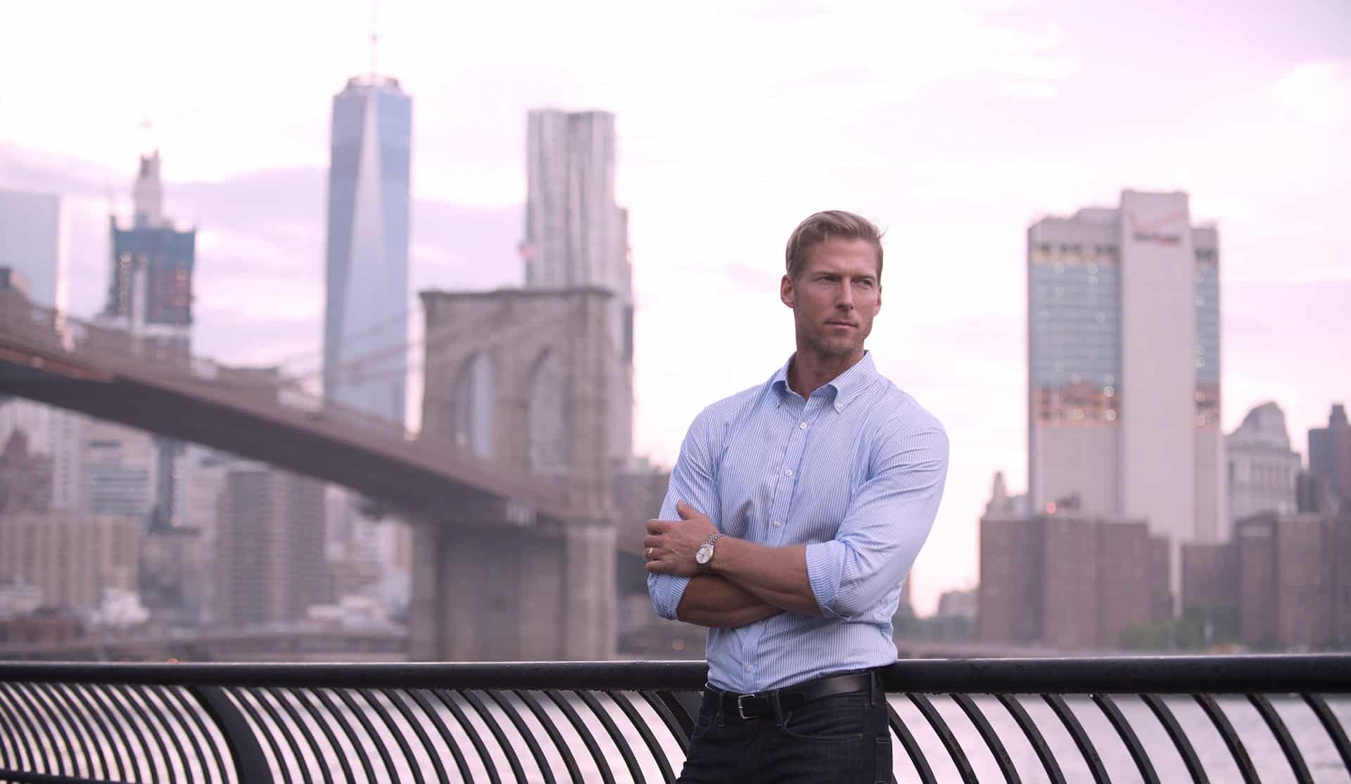 Modern businessman standing on Brooklyn Bridge overlooking Manhattan skyline during sunset, wearing a light blue dress shirt and wristwatch.