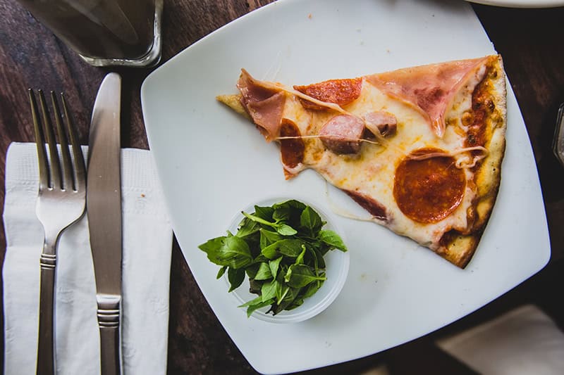 Thin crust pizza slice with ham, pepperoni, and cheese on a white plate, served with fresh basil garnish, set on a rustic wooden table. Perfect for pizza lovers and casual dining.