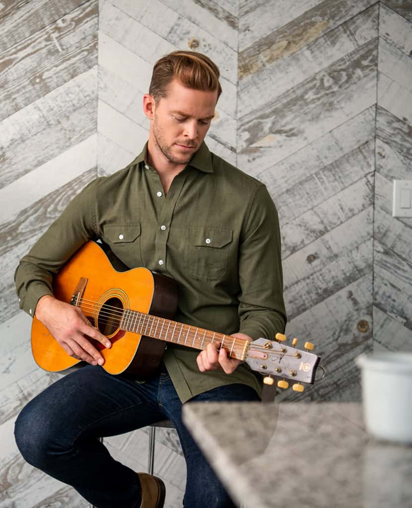 Relaxed man playing acoustic guitar in modern interior style, casual music performance, male musician, indoor musical setting, guitar player in cozy home environment, DeoVeritas music brand, lifestyle and entertainment image, high-quality music image, contemporary decor with wood accents, musical instrument, artistic expression, warm ambiance, professional music shoot, bohemian vibe, leisure activity, musical talent, creative hobby, tranquil music scene, home music studio, modern house decor with wooden wall panels.