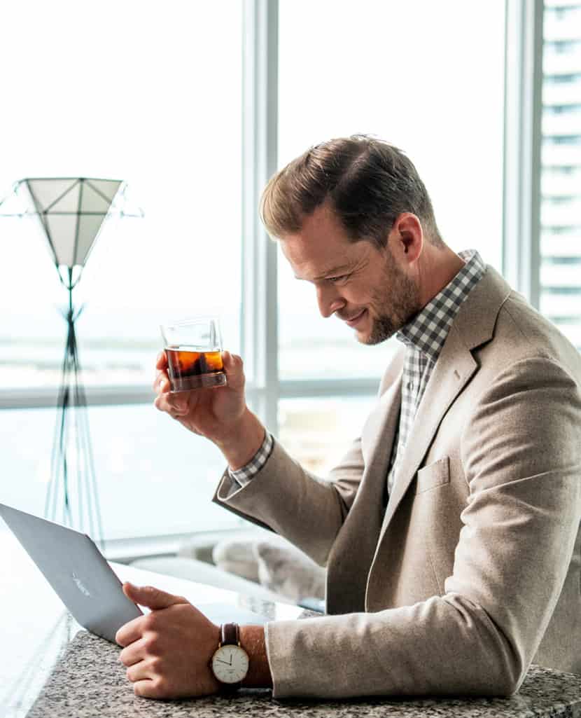 Man in a beige suit enjoying a glass of whiskey while working on a laptop in a modern office setting.