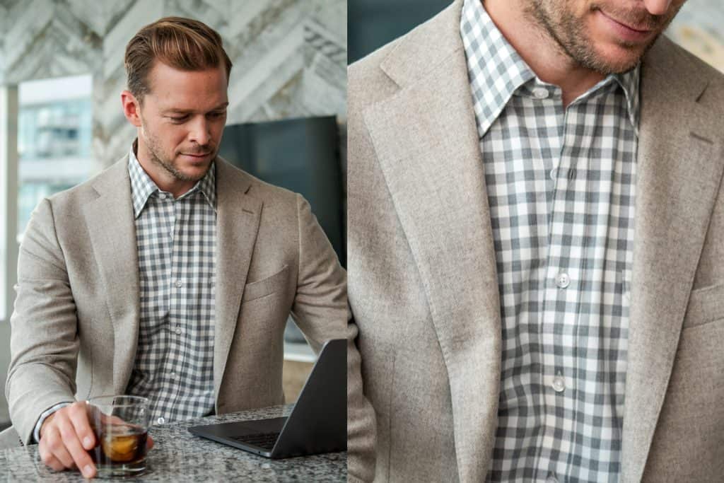 Elegant man in beige blazer and checkered shirt working on laptop, holding a glass of whiskey in a modern office setting.