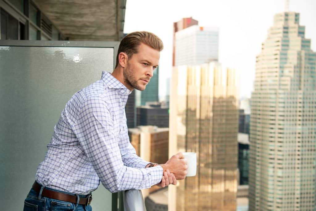 Modern businessman on a city balcony holding a white coffee mug, contemplating while enjoying a panoramic view of skyscrapers in the background.