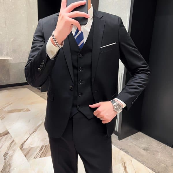 Elegant businessman in tailored black suit with white shirt and blue striped tie, taking a mirror selfie in a modern interior, accessorized with a silver watch and bracelet, showcasing professional attire.