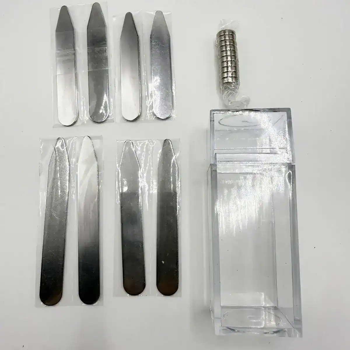 Precision metal and acrylic tools with small parts on a white background for personal care or grooming, featuring stainless steel blades, magnetic disks, and a clear acrylic display case.