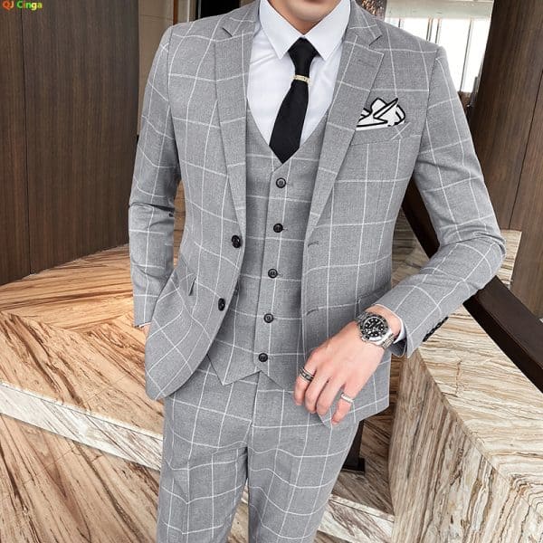 Sophisticated man wearing a tailored gray checkered suit, white shirt, black tie, and accessorized with a luxury watch, rings, and pocket square, standing indoors with modern wooden decor.